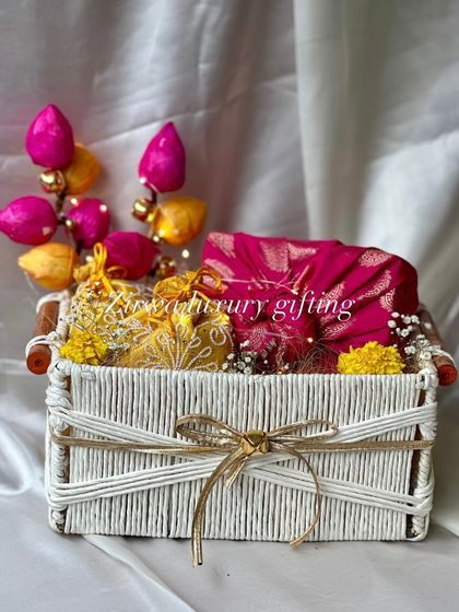Spreading the joy of Lohri with colourful hampers. This white basket is filled with bright pink and yellow items, including decorative potlis and a beautiful fabric piece.