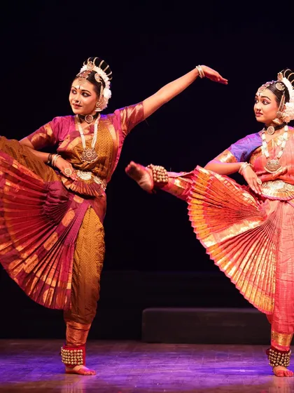 Two dancers in a dynamic pose with legs extended, their vibrant pleated costumes fanning out beautifully. A moment of pure artistry from the Sadhana festival.