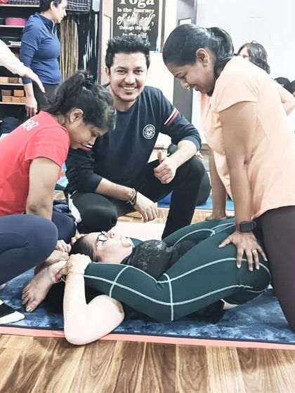 An instructor smiles as students successfully assist a fellow yogi in a deep hip-opening posture.