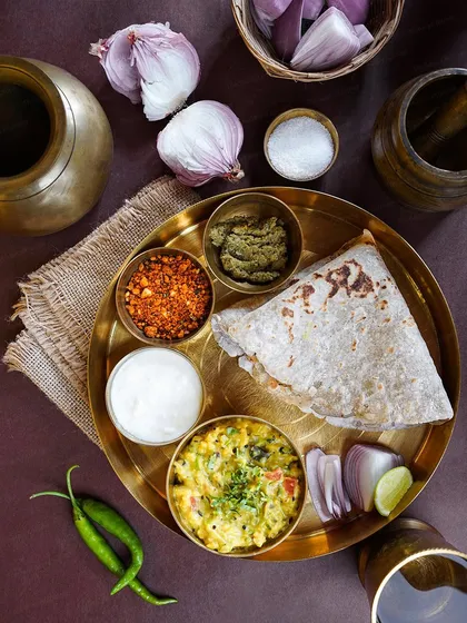 A flatlay of a traditional Maharashtrian meal of Jhunka Bhakar with thecha and yogurt. The composition with raw ingredients like garlic and onions highlights the dish's simple, rustic origins.