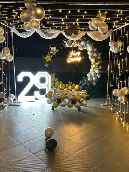 A 29th birthday party on a terrace. This decoration includes a canopy of fairy lights, a circular balloon arch in black and silver, a neon sign, and marquee numbers.