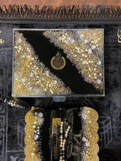 A top-down view of my trending black and gold Islamic gift set. This luxurious collection includes a personalized prayer mat, a resin Quran box, and a matching Quran holder, all heavily embellished for a grand look.