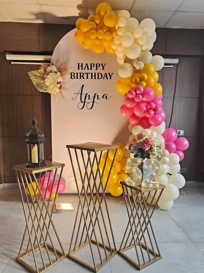 A vibrant and personalized setup for a 52nd birthday celebration. This features a custom backdrop with the name 'Appa', a colorful balloon garland in yellow and pink, and stylish pedestals for the cake and decor accents.