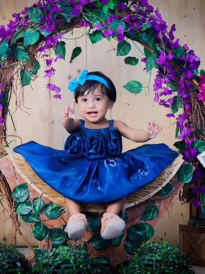 A one-year-old girl in a beautiful blue dress, sitting in a basket adorned with a wreath of purple flowers. A classic and elegant milestone portrait.