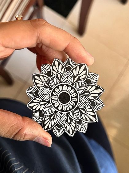 A small, hand-held mandala cutout. The intricate black and white pattern is shaped like a blooming flower, perfect for use as a decorative element or a unique bookmark.