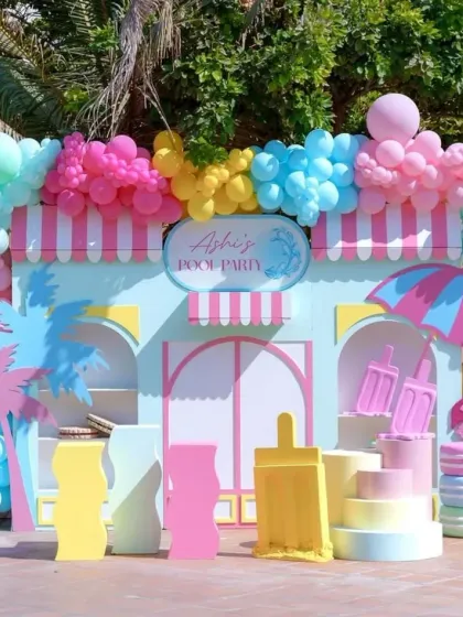 A vibrant and colorful pool party setup, perfect for a kids' summer birthday. The backdrop is designed like a fun beach house with a massive balloon garland.