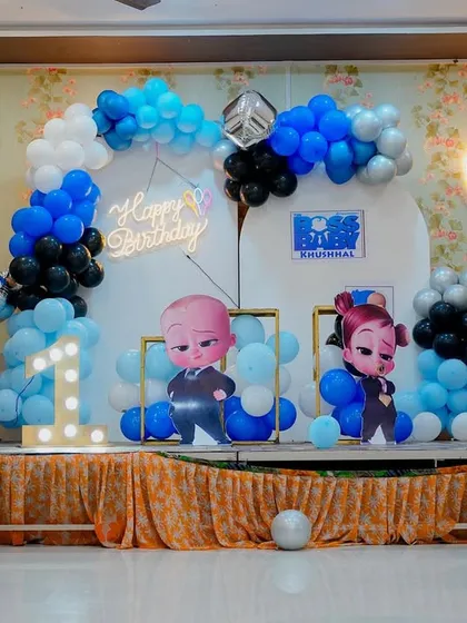 A Boss Baby themed first birthday stage setup. The decor includes a house-shaped backdrop, character cutouts, a light-up number '1', and balloon columns in a blue, black, and white color scheme.
