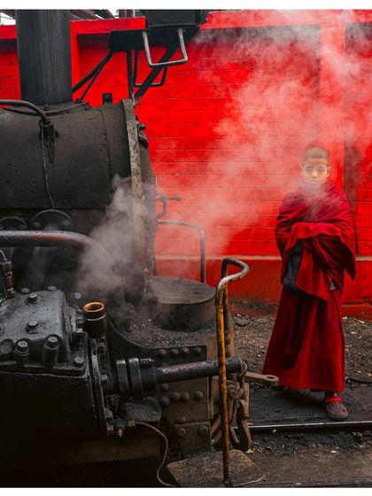 A young Buddhist monk stands in front of a steam engine in Darjeeling. The contrast of his red robes against the red wall and the steam creates a powerful and cinematic image.