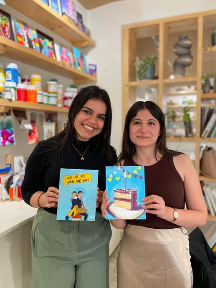 Two friends displaying their fun and quirky paintings, one of a birthday cake and the other a pop-culture reference.
