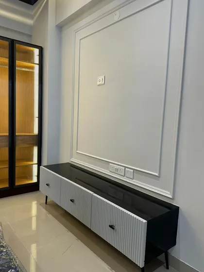 A minimalist TV console with a fluted white facade and a contrasting black top. This piece is set against a wall with simple, elegant moulding, creating a clean and contemporary look. The glass-fronted wardrobe nearby adds to the modern aesthetic.