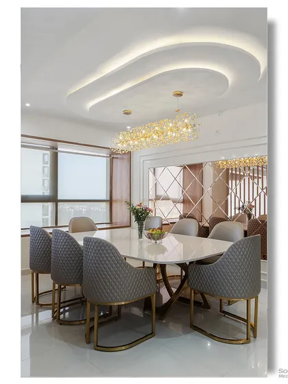 A luxurious dining room featuring quilted grey chairs, a marble table, and a stunning linear crystal chandelier. The curved ceiling detail and mirrored wall add layers of sophistication and glamour.