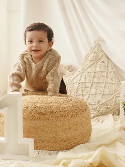 An adorable baby boy flashes a huge smile while posing on a jute pouf for his first birthday pictures.