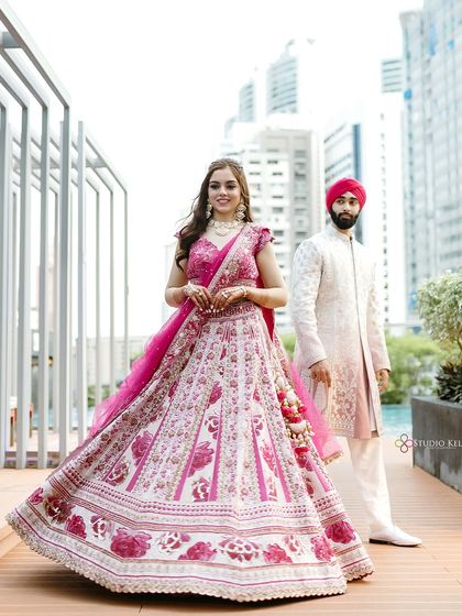 A stylish portrait of the bride twirling in her vibrant pink floral lehenga, with the groom and the modern Bangkok cityscape behind her.