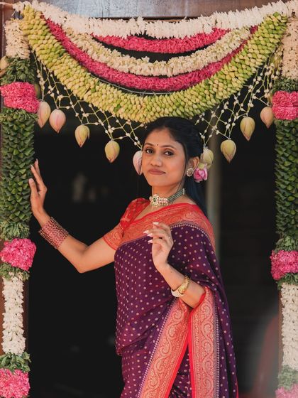 The bride stands at a doorway beautifully decorated with fresh flowers and mango leaves, a traditional symbol of welcome and auspiciousness for the "Chapra Pooje".