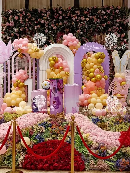 A close-up view of a butterfly-themed first birthday stage. This detailed shot highlights the lush floral base, pastel and gold balloon cascades, and intricate butterfly cutouts, all roped off with elegant red velvet ropes.
