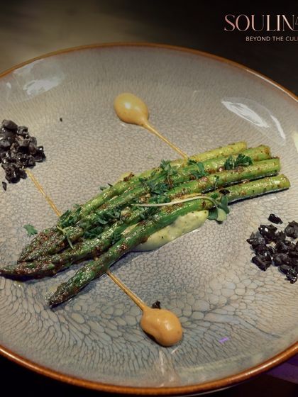 A plate of perfectly grilled asparagus, artistically arranged with dollops of sauce and a sprinkle of black olive crumble. We turn simple, fresh ingredients into a sophisticated culinary experience.
