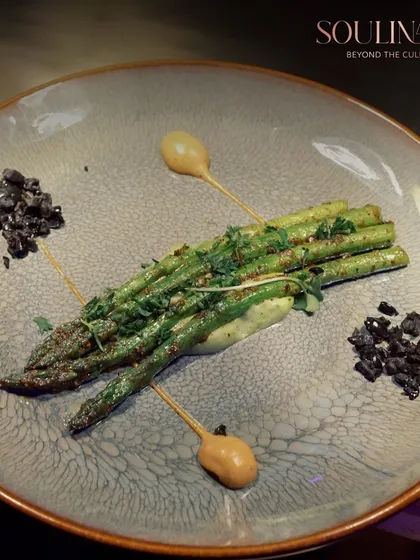 A plate of perfectly grilled asparagus, artistically arranged with dollops of sauce and a sprinkle of black olive crumble. We turn simple, fresh ingredients into a sophisticated culinary experience.