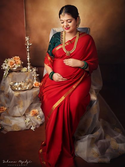 A serene and graceful solo portrait. The mom-to-be sits peacefully, her hands on her belly, in a setting filled with traditional Indian decor elements.