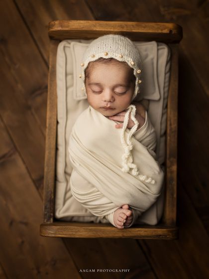 A sweet new blossom of humanity. This classic overhead shot shows a peacefully sleeping newborn, wrapped in neutral tones and wearing a delicate pearl-adorned bonnet in a tiny wooden bed.