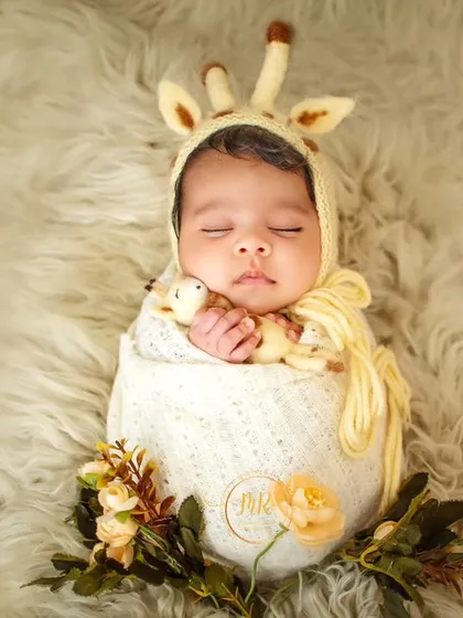 A sleeping newborn wearing an adorable giraffe-themed bonnet, swaddled in a cream-colored wrap on a soft, furry surface.