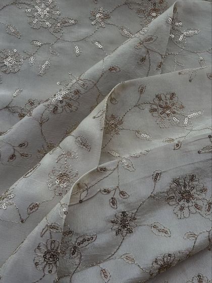 This grey embroidered tissue fabric features a delicate floral vine pattern with sequin accents. It's a sophisticated and elegant choice for a saree or suit set.