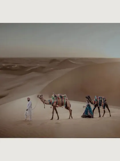 A cinematic wide shot capturing a couple's journey through the vast desert dunes of Rajasthan. This image highlights our ability to create grand, story-driven visuals for adventurous couples, complete with elements like traditionally adorned camels.