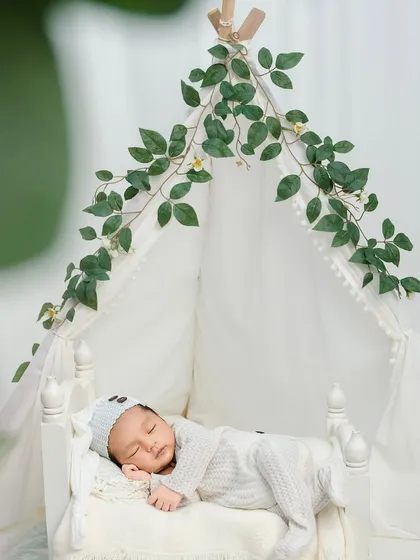 A newborn sleeps soundly on a miniature bed under a leafy canopy. This whimsical setup adds a touch of nature and fairytale magic to a classic newborn portrait.