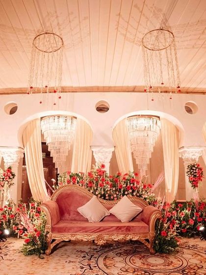 An opulent indoor reception stage, with a vintage pink sofa surrounded by a garden of red flowers, set against a backdrop of elegant arches and cascading crystal chandeliers.
