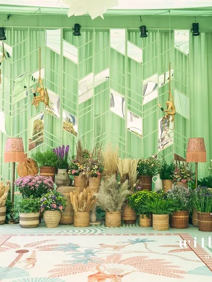 A wide shot of 'The Conservatory' setup, featuring a custom photo-op area with a mirrored wall and playful monkey light fixtures. The space is filled with potted plants and flowers, blurring the line between indoors and out.