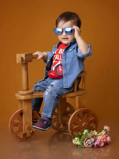 A fun and stylish studio portrait of a young boy for his pre-birthday photoshoot. He is dressed in a denim jacket and sunglasses, posing confidently on a wooden tricycle prop.