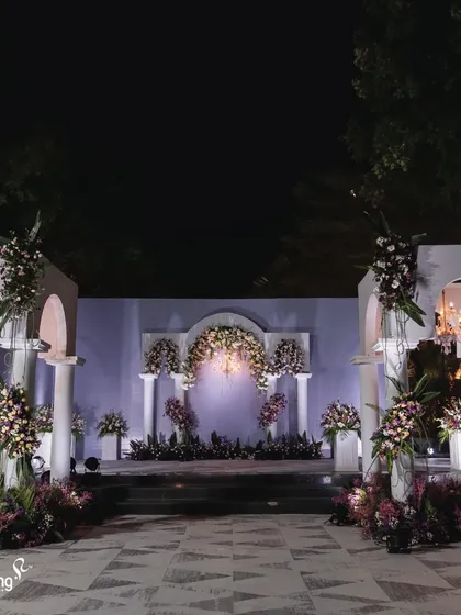 A grand, white architectural stage for a reception. The Roman-style pillars and arches, adorned with purple and white flowers, create a majestic and dramatic setting.