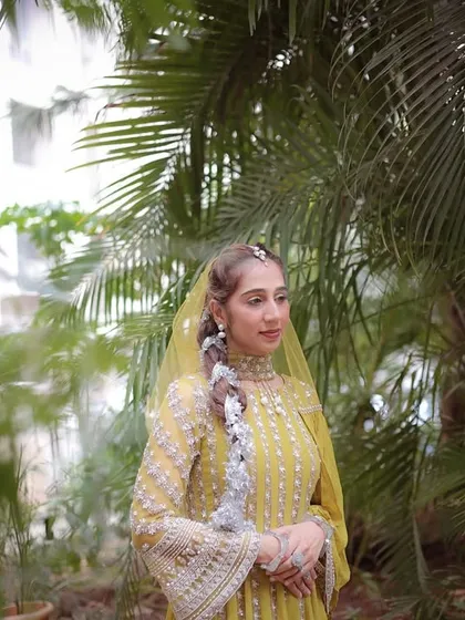 A full-length portrait of the bride amidst tropical foliage. The vibrant green leaves provide a stunning natural backdrop for her traditional yellow dress and intricate braid.