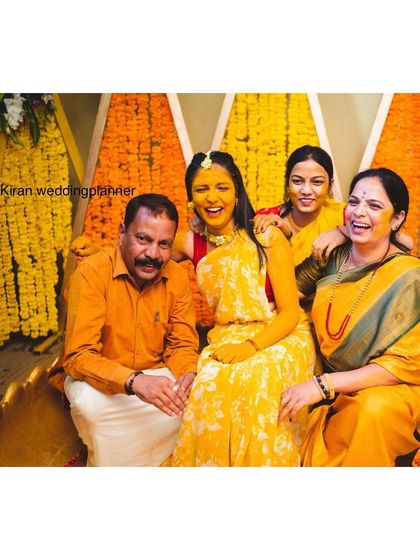 A happy family photo from a Haldi ceremony, showing everyone dressed in yellow against a backdrop of marigold and orange flowers.
