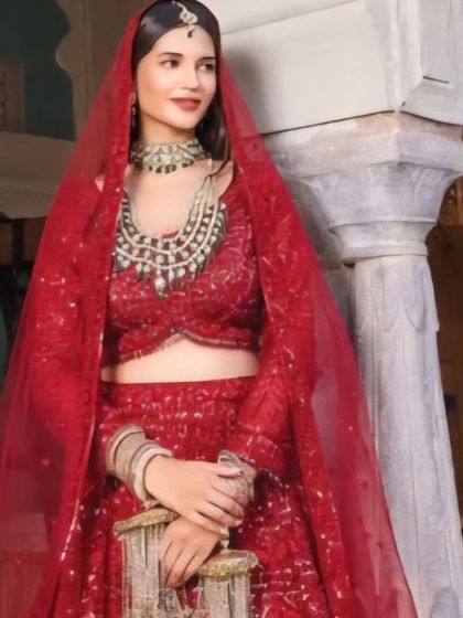 A close-up of a bride in a traditional red lehenga, complete with a sheer red veil. The heavy, layered necklace and sparkling details on the outfit make this a classic and timeless bridal look.