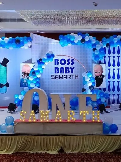 A grand Boss Baby themed stage decoration perfect for a first birthday party. This large-scale setup features multiple themed backdrops, extensive blue and white balloon arches, character cutouts, and illuminated marquee letters spelling out the birthday boy's name.