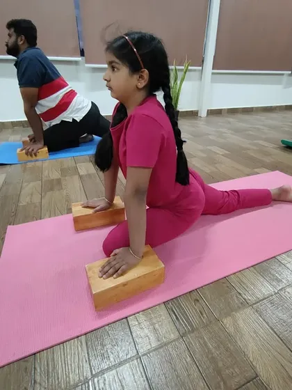 A young student uses blocks to support her in Rajakapotasana (Pigeon Pose). Using props is essential for kids to learn correct alignment and safely work on flexibility without forcing anything.