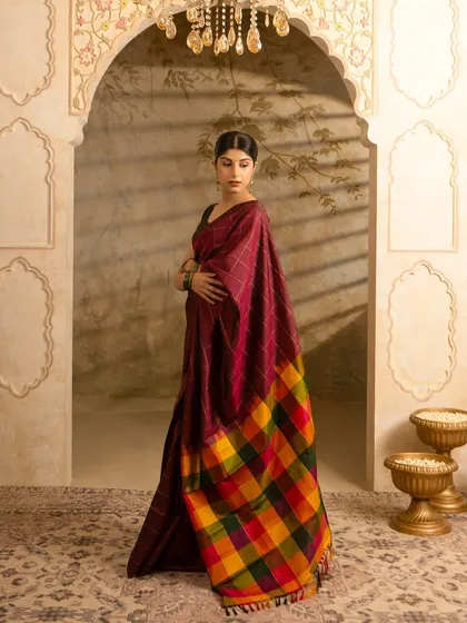 An editorial-style photograph for Kumaran Silks, showcasing a traditional saree in a grand, architectural setting. The lighting and composition are designed to highlight the fabric's drape, rich color, and the intricate pattern of the pallu, creating an aspirational look for fashion brands.
