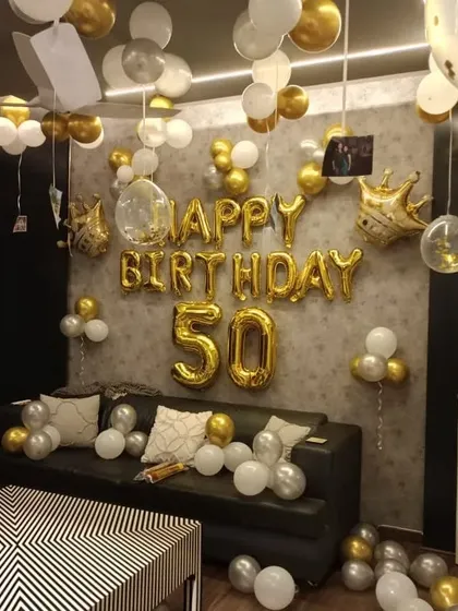 A 50th birthday celebration at home with gold and white balloons filling the room. Gold foil balloons spell out 'HAPPY BIRTHDAY 50'.