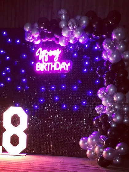 An 18th birthday setup with a touch of dark glamour. We used a black sequin wall with purple pixel lights, a "Happy Birthday" neon sign, and a black and silver balloon garland to create a cool and sophisticated vibe for this milestone event.