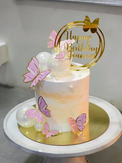 A delicate butterfly-themed cake for little Joanna. The cake has a soft watercolor finish and is adorned with shimmering pearl-like spheres and beautiful pink butterflies fluttering around it.