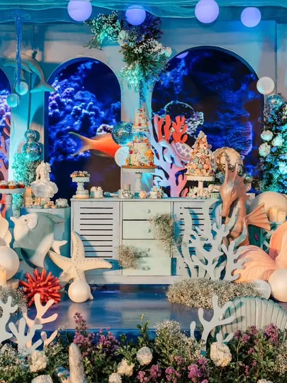The dessert table is a masterpiece of underwater design, set against a digital backdrop of a coral reef. The table is surrounded by custom props of seashells, starfish, and coral.