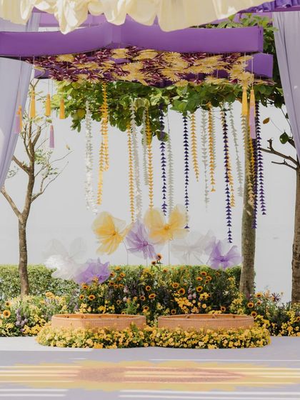 The main ceremony setup for the pastel Haldi. The seating for the couple is framed by a canopy with cascading floral strings and large, whimsical butterfly bows, creating a dreamy and romantic focal point.