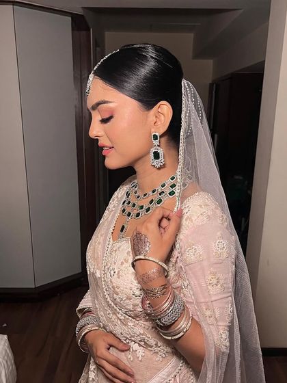 A sleek and elegant bridal bun, perfect for a Nikah ceremony. The clean lines of the hairstyle beautifully complement the delicate veil and emerald jewelry.