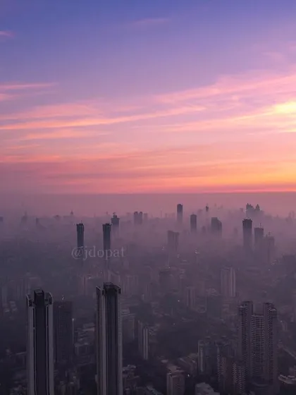 A high-altitude drone shot shows the clean, pink and purple sky above a thick layer of smog covering Mumbai.