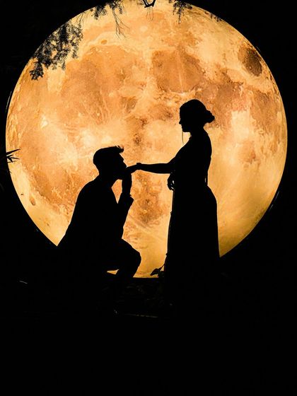 A stunning silhouette of a groom-to-be kneeling and kissing his fiancée's hand against a large, glowing moon prop, creating a perfectly romantic pre-wedding scene.