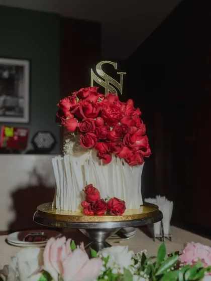 A dramatic and romantic two-tiered wedding cake. This design features white chocolate shards, a cascade of fresh red roses, and a custom gold initial topper for the couple, 'S' and 'N'.