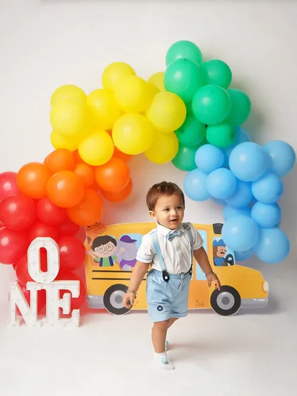 A one-year-old boy stands proudly in front of a 'Wheels on the Bus' themed backdrop with a rainbow balloon arch. This is a fun and colorful theme for a first birthday.