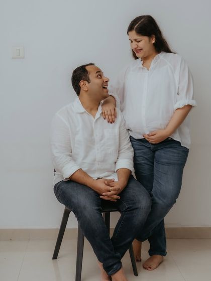 A full-length portrait showing the couple's relaxed dynamic during their home photoshoot. The simple posing against a plain wall keeps the focus entirely on their connection and anticipation.