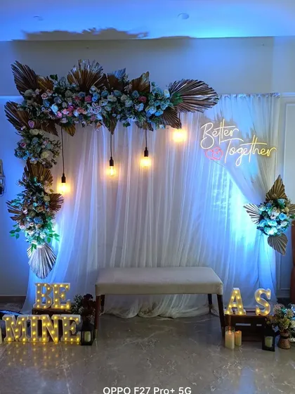 A complete view of a home engagement setup. It includes a floral arch with dried palms, a "Better Together" sign, "Be Mine" marquee letters, and a bench for seating.