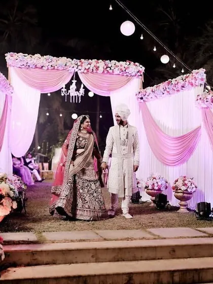 A couple makes their grand entrance against a backdrop of pink and white drapes and floral arrangements. The stage is designed to be both elegant and welcoming for the wedding reception.